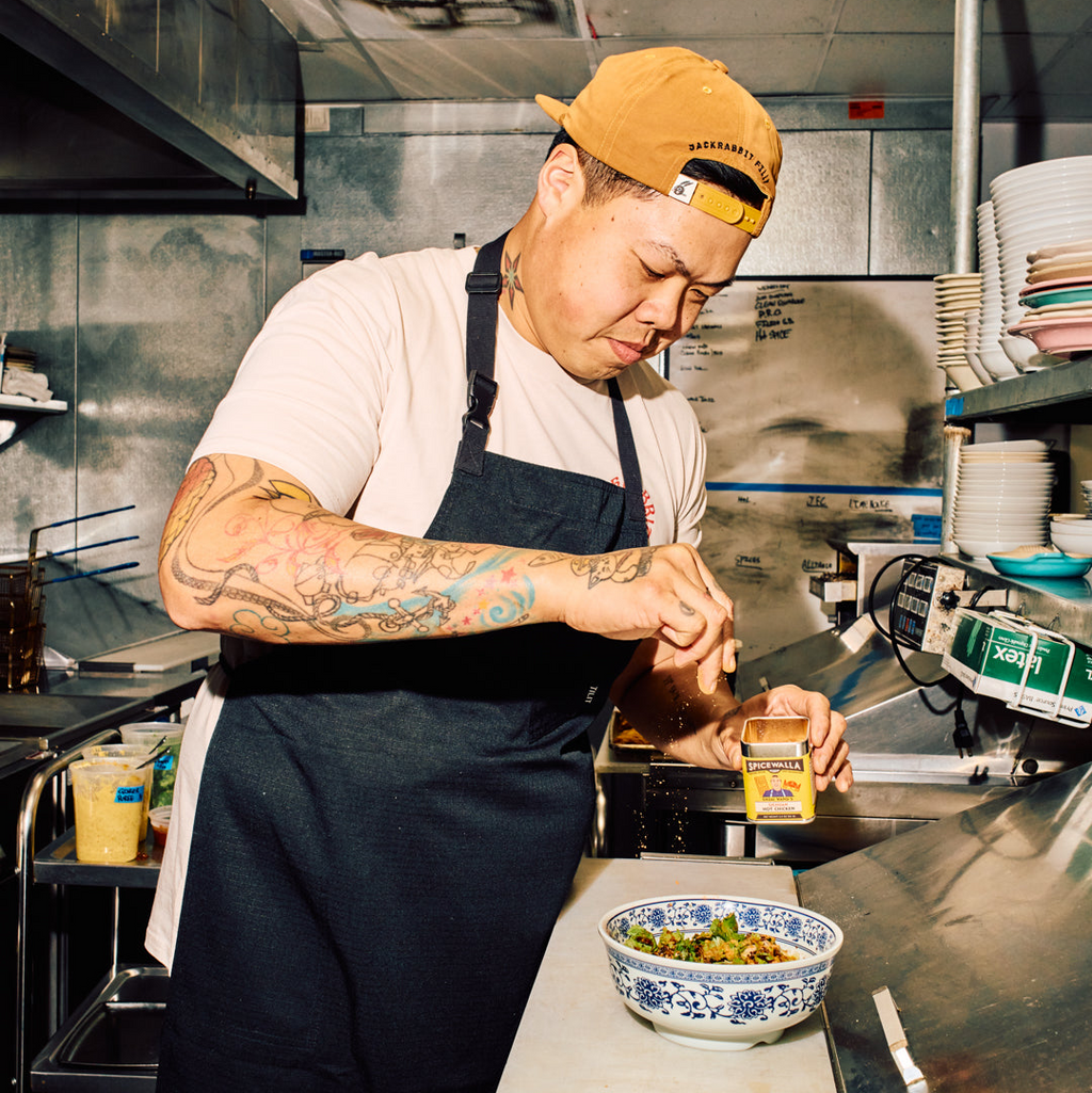 Person in a kitchen wearing an apron and yellow cap, preparing food.